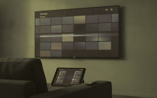 A TV showing a programme guide grid with a tablet and phone showing channel lists on a sofa