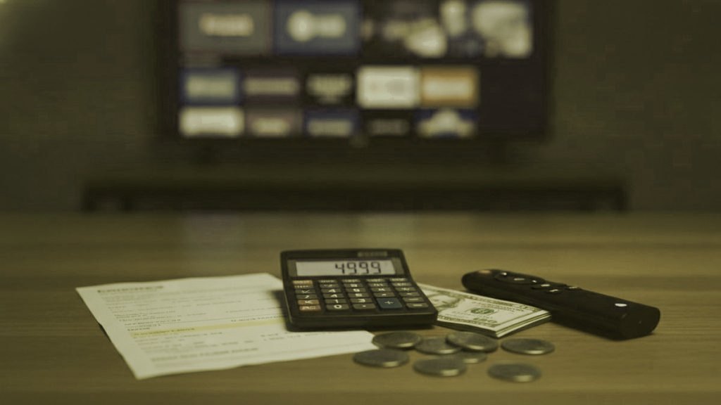 A calculator, dollar bills, coins, a streaming stick and a remote on a table in front of a TV