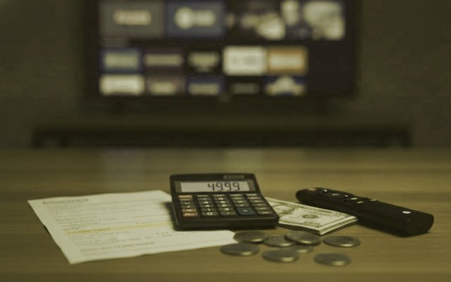 A calculator, dollar bills, coins, a streaming stick and a remote on a table in front of a TV